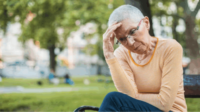 A lady with cropped gray hair and glasses puts her hand to her forehead, looking upset